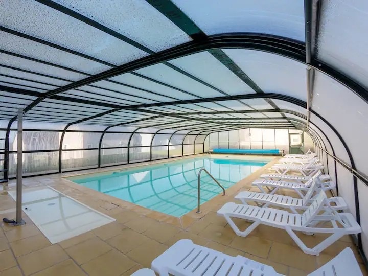 An indoor swimming pool is enclosed under a translucent roof, allowing natural light to fill the space. White lounge chairs are arranged neatly along the poolside. The clear pool water reflects the surrounding structures, and a shallow area is visible at one end.