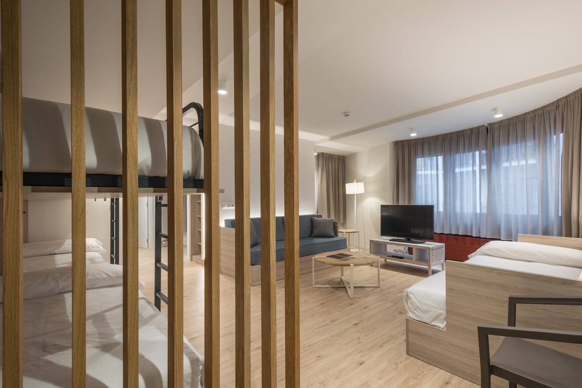 An open-plan living space features a cozy layout with a bunk bed and two separate sleeping areas, each with spacious beds. A comfortable sofa is positioned next to a television, while soft natural light filters through large windows with sheer curtains.