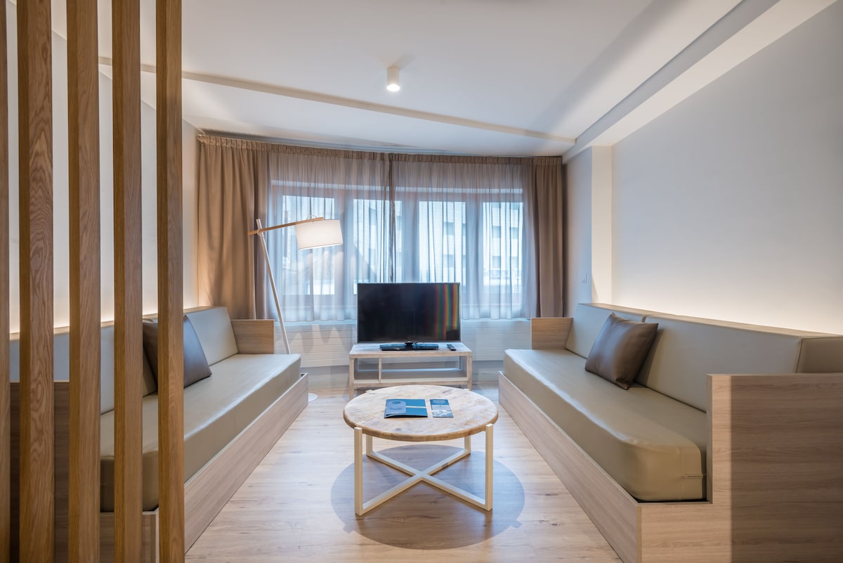 A modern living area is presented with soft beige seating along two walls. A round coffee table sits centrally, complemented by a television mounted on a wall. Natural light enters through wide windows, adorned with sheer curtains, creating a bright and airy environment.
