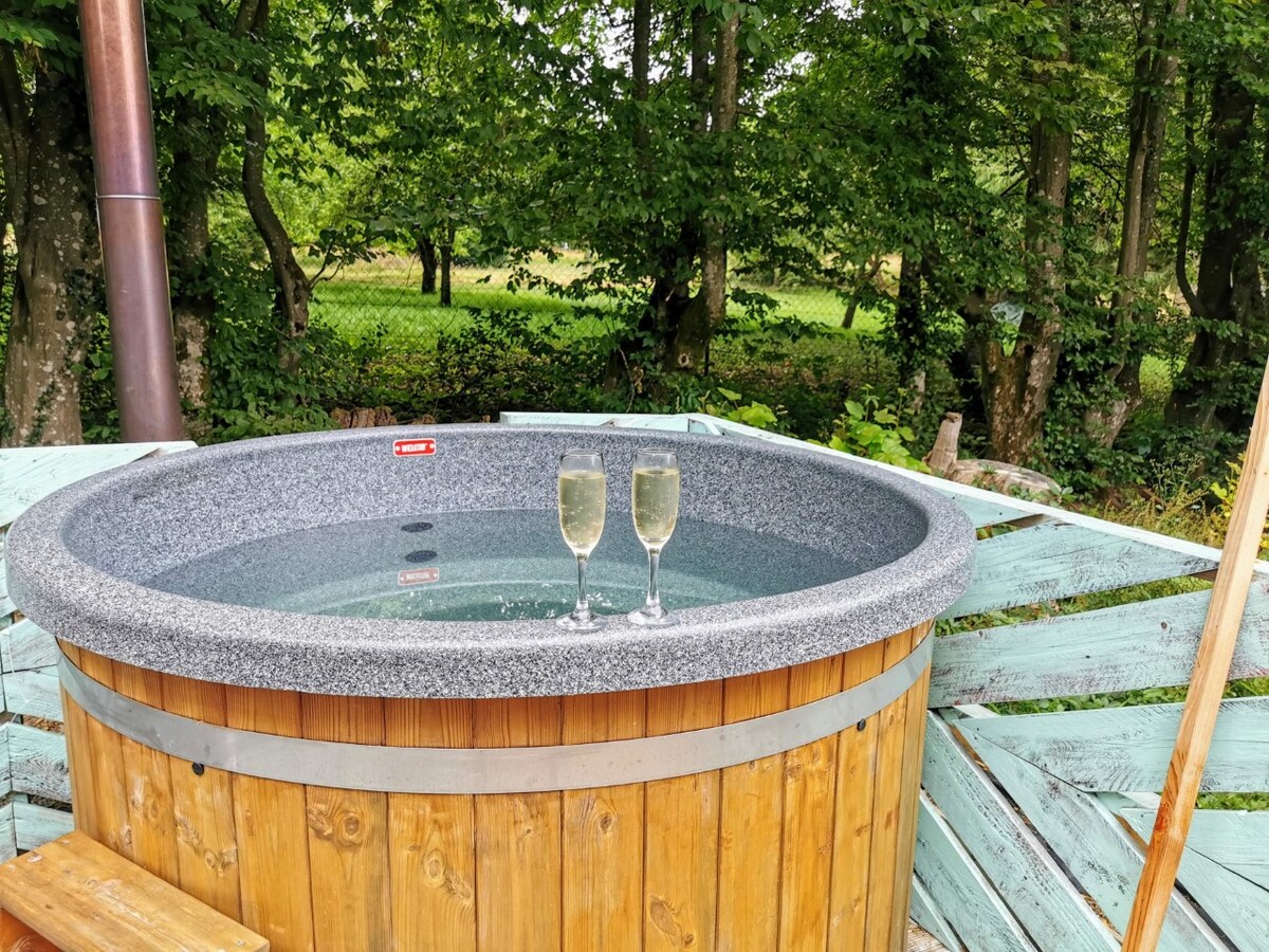 A wooden hot tub is situated on a deck surrounded by trees, providing a serene outdoor setting. Two glasses of sparkling beverage are placed on the rim of the tub, reflecting the natural surroundings.