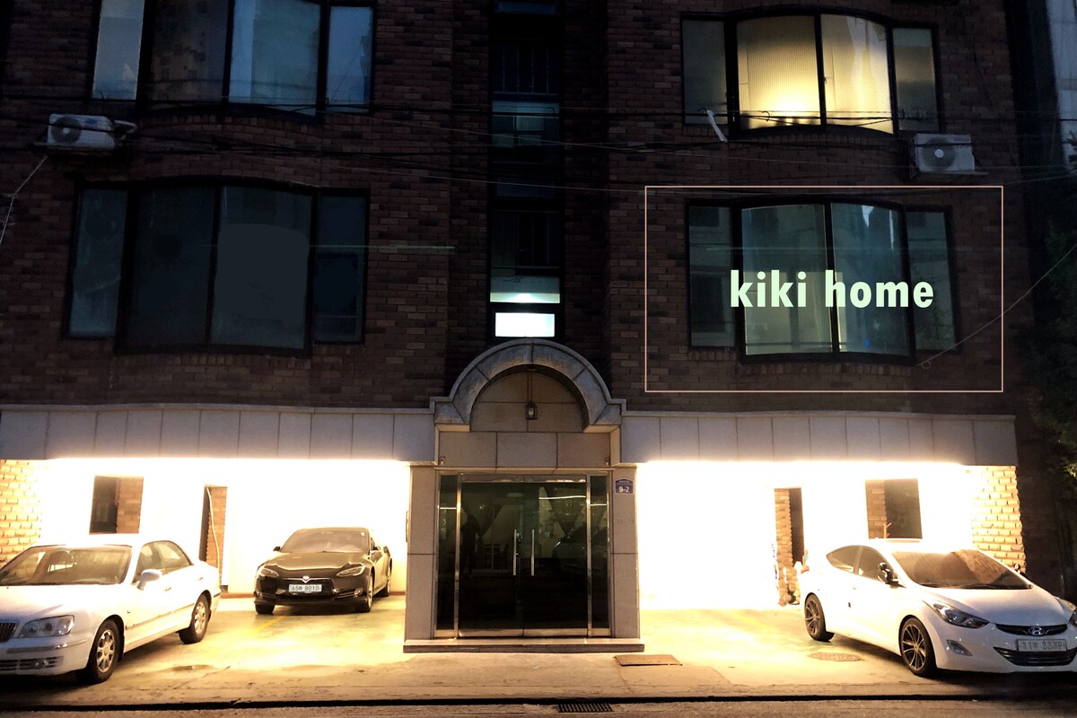 The exterior of the building is shown at night, featuring parking spaces for multiple vehicles. The entrance is highlighted by soft lighting, and a sign displaying 'kiki home' is visible above the doorway. Large windows reflect light, contributing to the welcoming appearance.