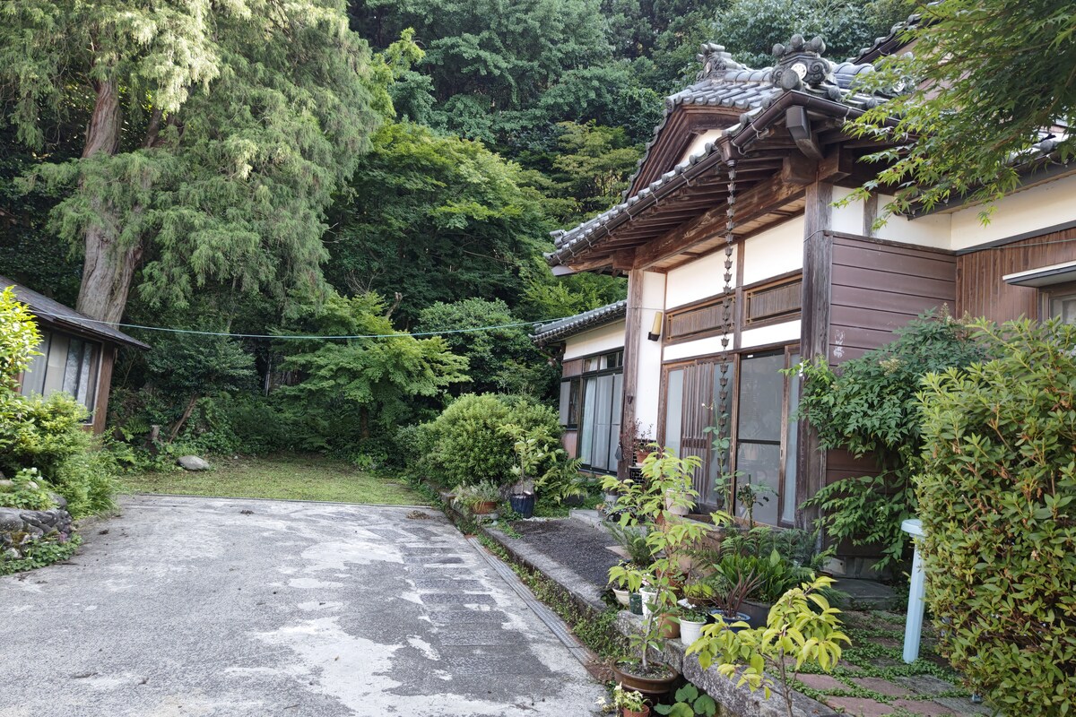 The exterior of the property features traditional Japanese architecture, with wooden beams and a tiled roof. Lush greenery surrounds the entrance, and a pathway leads up to the main building. The landscaped area includes various plants and decorative elements.