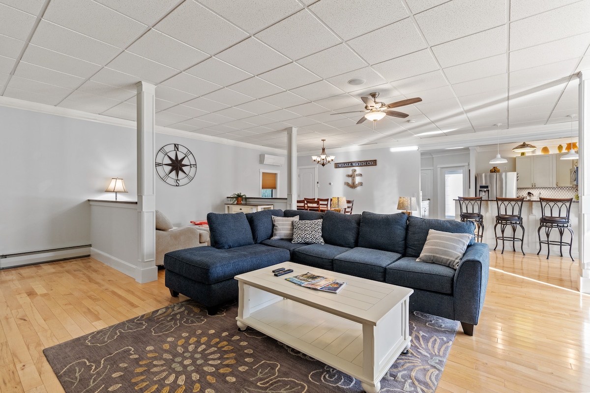 The living area features a spacious sectional couch in a deep blue hue, complemented by a light coffee table. Hardwood floors add warmth, and a large area rug outlines the seating area. To the back, a dining table is visible, along with bar seating near the kitchen.
