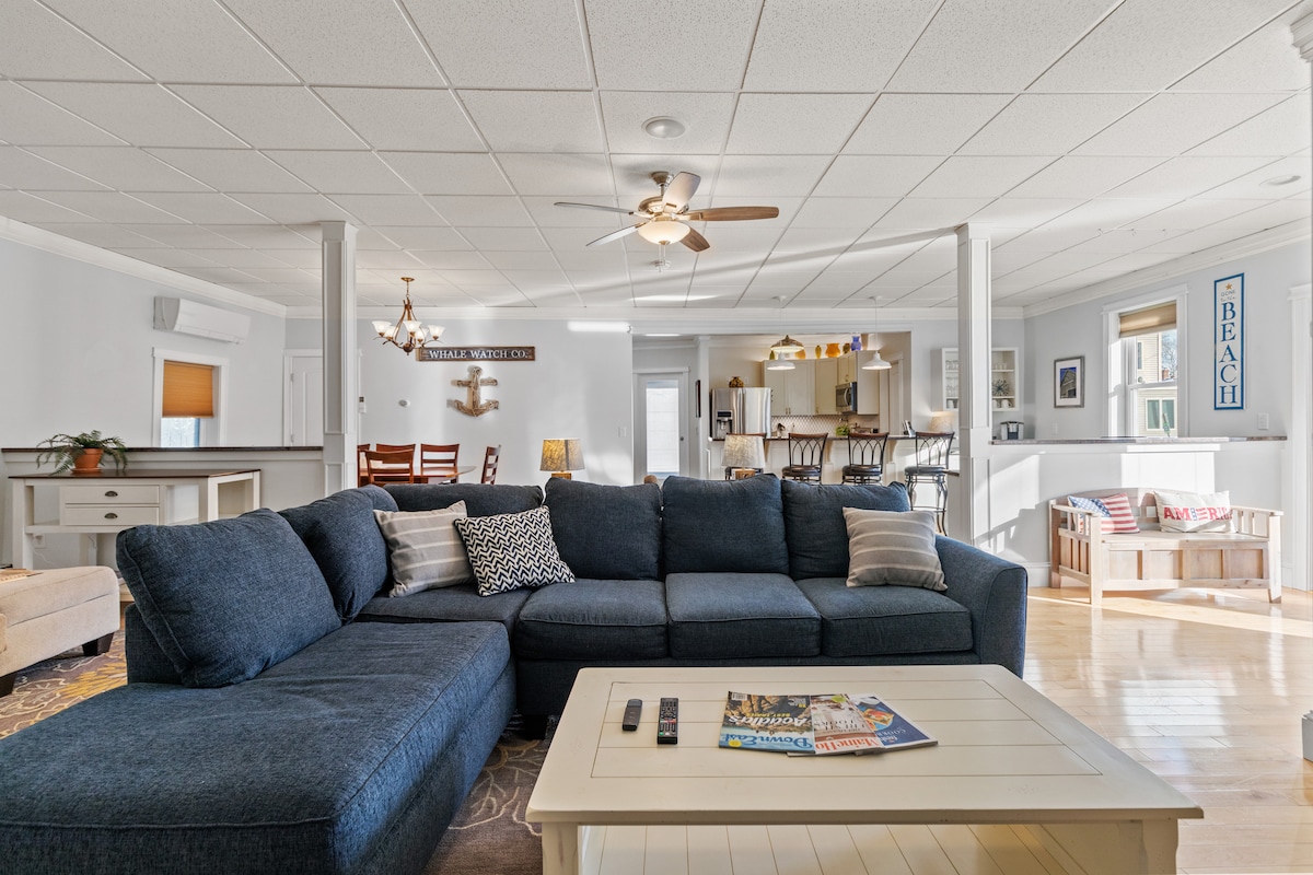 An open-concept living room features a large sectional couch in a navy hue, complemented by a coffee table and a decorative magazine. Light-colored walls and ceiling tiles enhance the brightness of the space, while the kitchen area is visible in the background, showcasing bar seating.