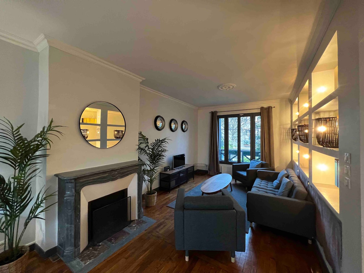 A light-filled living area features two large windows with views overlooking the garden terrace. A comfortable gray sofa and an armchair are positioned around a circular coffee table. Decorative mirrors adorn the walls and a sleek fireplace adds a classic touch.