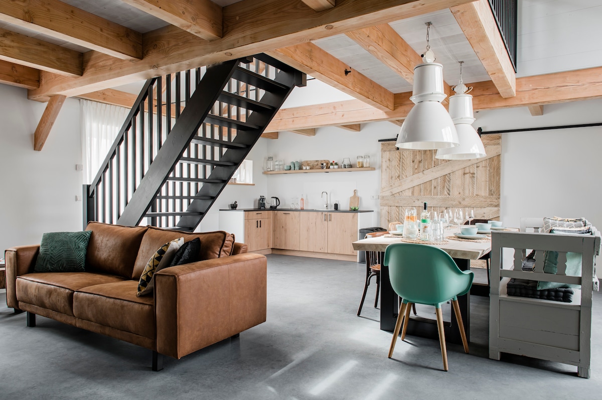 An open-concept living area features a comfortable brown sofa, a dining table set for six, and a fully equipped kitchen. Large pendant lights hang above, illuminating the space. Natural wood beams and a black staircase offer a modern rustic aesthetic.