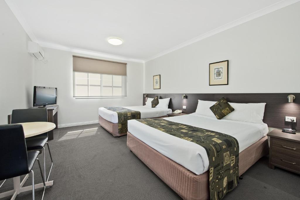 A compact room is furnished with two queen beds, each featuring decorative throw pillows. A small round table and two chairs are positioned beside a flat-screen television. Natural light enters through a window with a neutral-toned blind, creating a functional and welcoming space.
