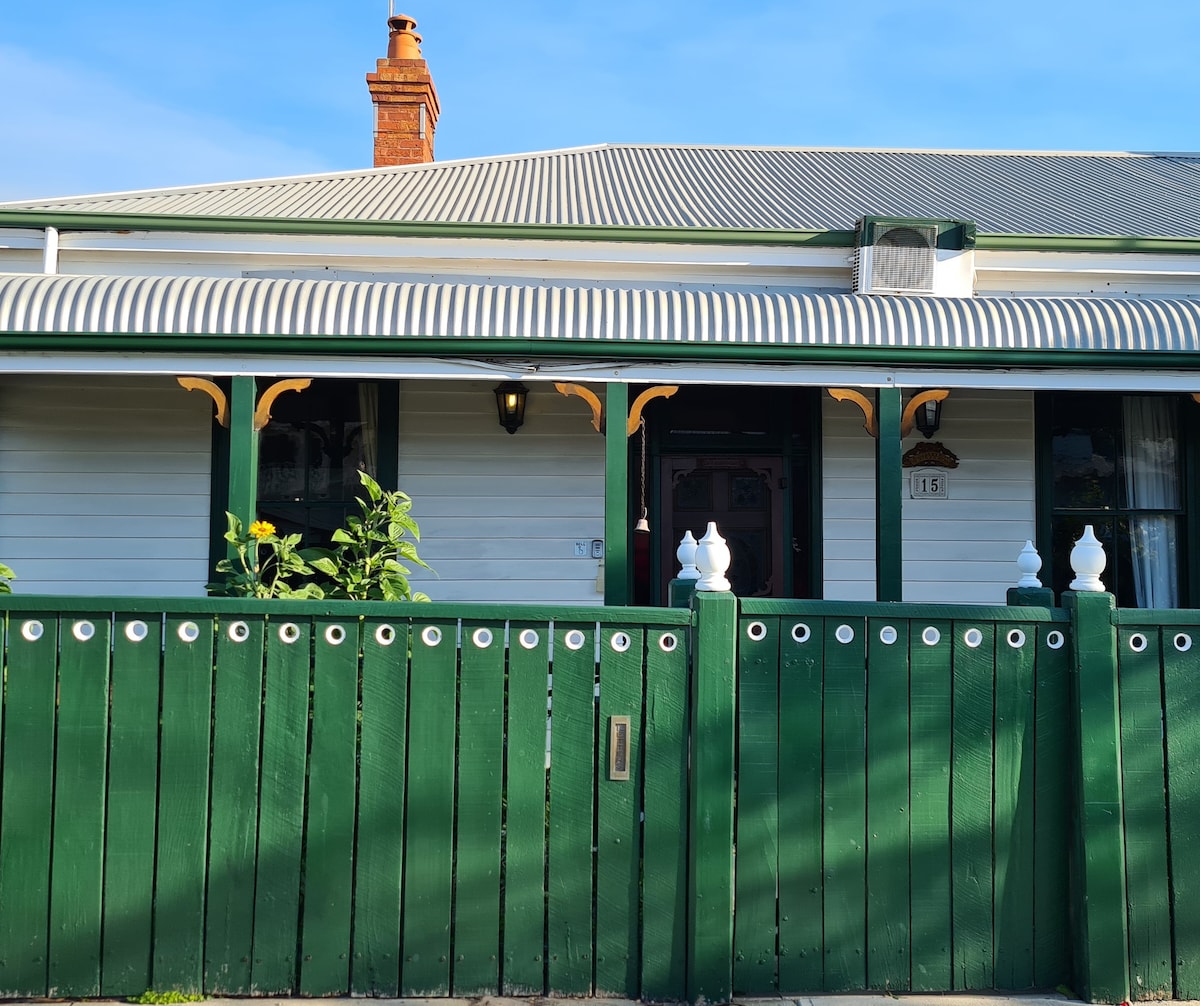 A charming façade of a historic house displays a green picket fence accented with white circular details. The entrance is framed by decorative woodwork and is complemented by a chimney rising from the roof, with plants adding a touch of greenery.