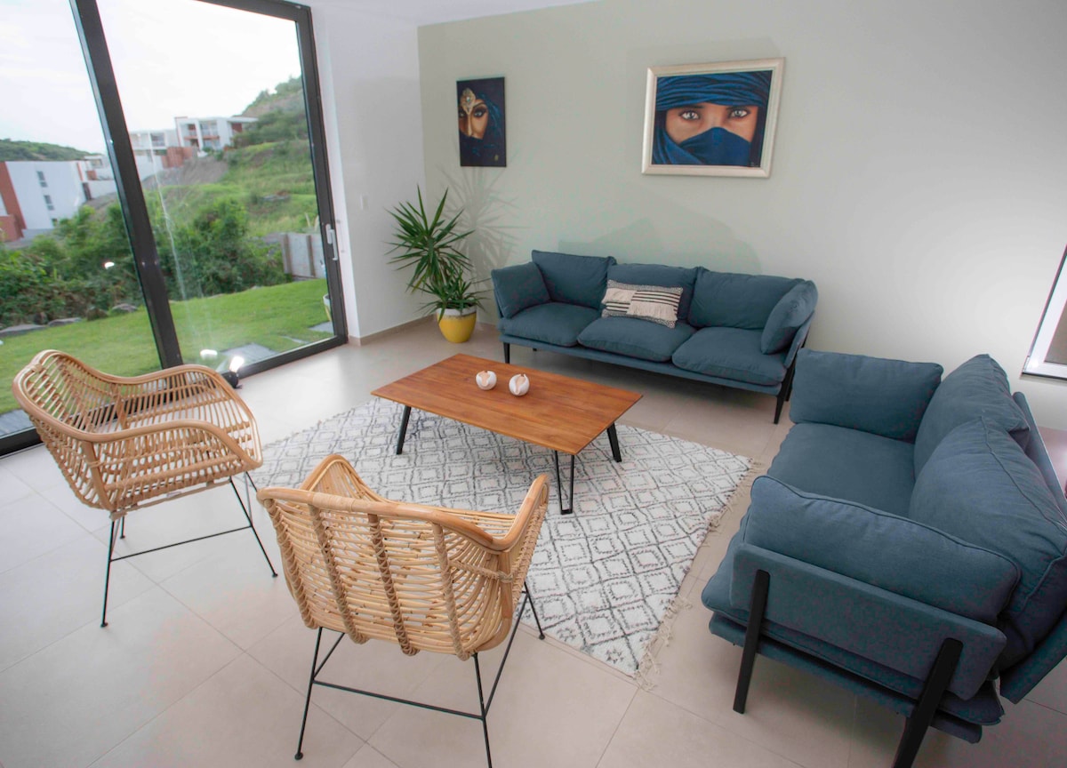A modern living area features a blue sofa and two rattan chairs, arranged around a wooden coffee table. A patterned area rug is placed on the tiled floor. Large glass doors allow natural light to fill the space and provide views of the green surroundings.