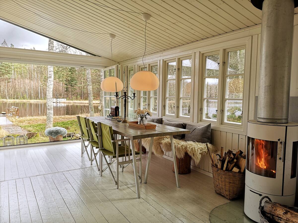A spacious dining area features a long table surrounded by light-colored chairs, illuminated by two pendant lights. Large windows create a bright atmosphere and offer views of the serene lake and surrounding forest. A wood-burning stove adds warmth, with a basket of firewood nearby.
