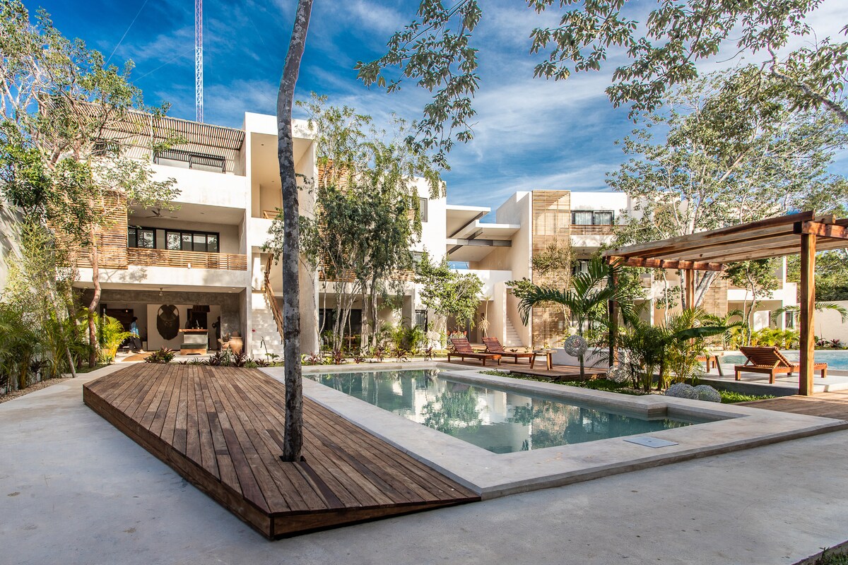 A serene common area is depicted, featuring two pools surrounded by lush greenery. Wooden decking leads to a shallow pool, while comfortable lounge chairs are visible. The architecture showcases a blend of modern design and natural elements, enhancing the tranquil atmosphere.