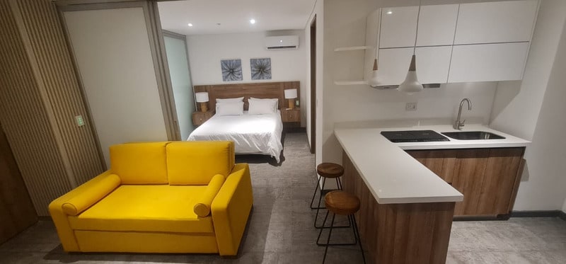 The image displays a contemporary studio apartment featuring a yellow sofa in the living area. A spacious bed with crisp white linens is positioned in the background. An equipped kitchen with modern cabinetry and a breakfast bar is visible, along with two stools for seating.