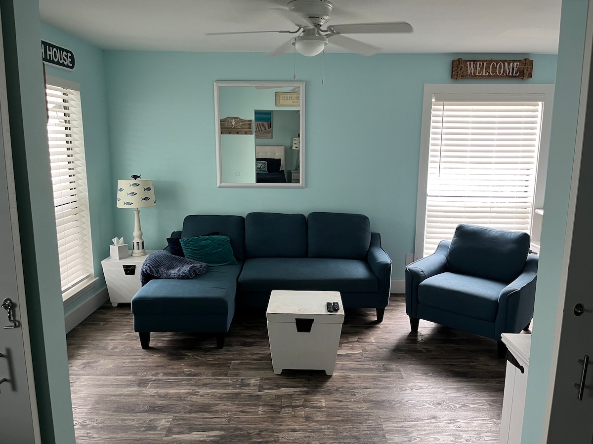 A light blue living area is presented, featuring a sectional sofa with a matching armchair. A coffee table and a lamp are included. Natural light filters through the white blinds, enhancing the relaxed ambiance. A welcome sign is displayed on the wall, contributing to the cozy environment.