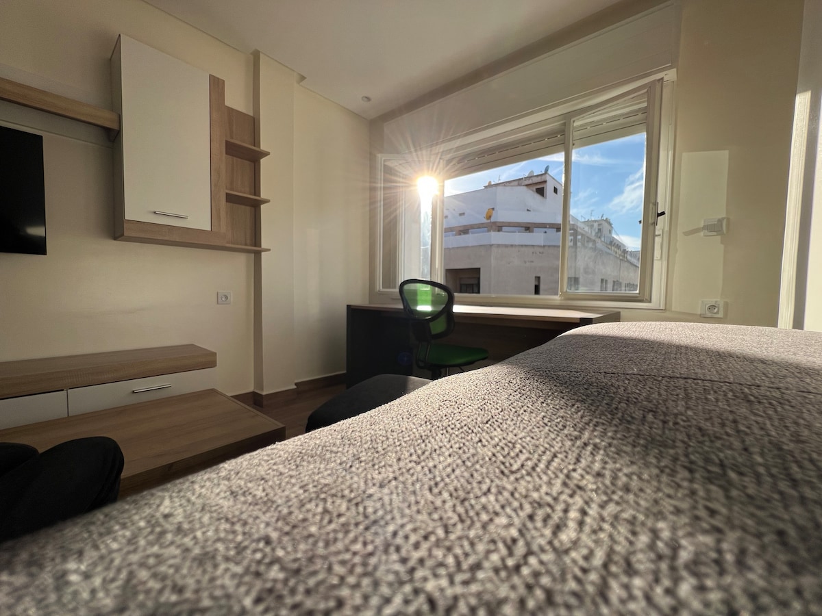 A well-lit room features a large window with sunlight streaming in. A desk with a green chair is positioned adjacent to a cozy bed, while a wall-mounted television and shelving units are visible. The neutral color palette creates a calm environment.