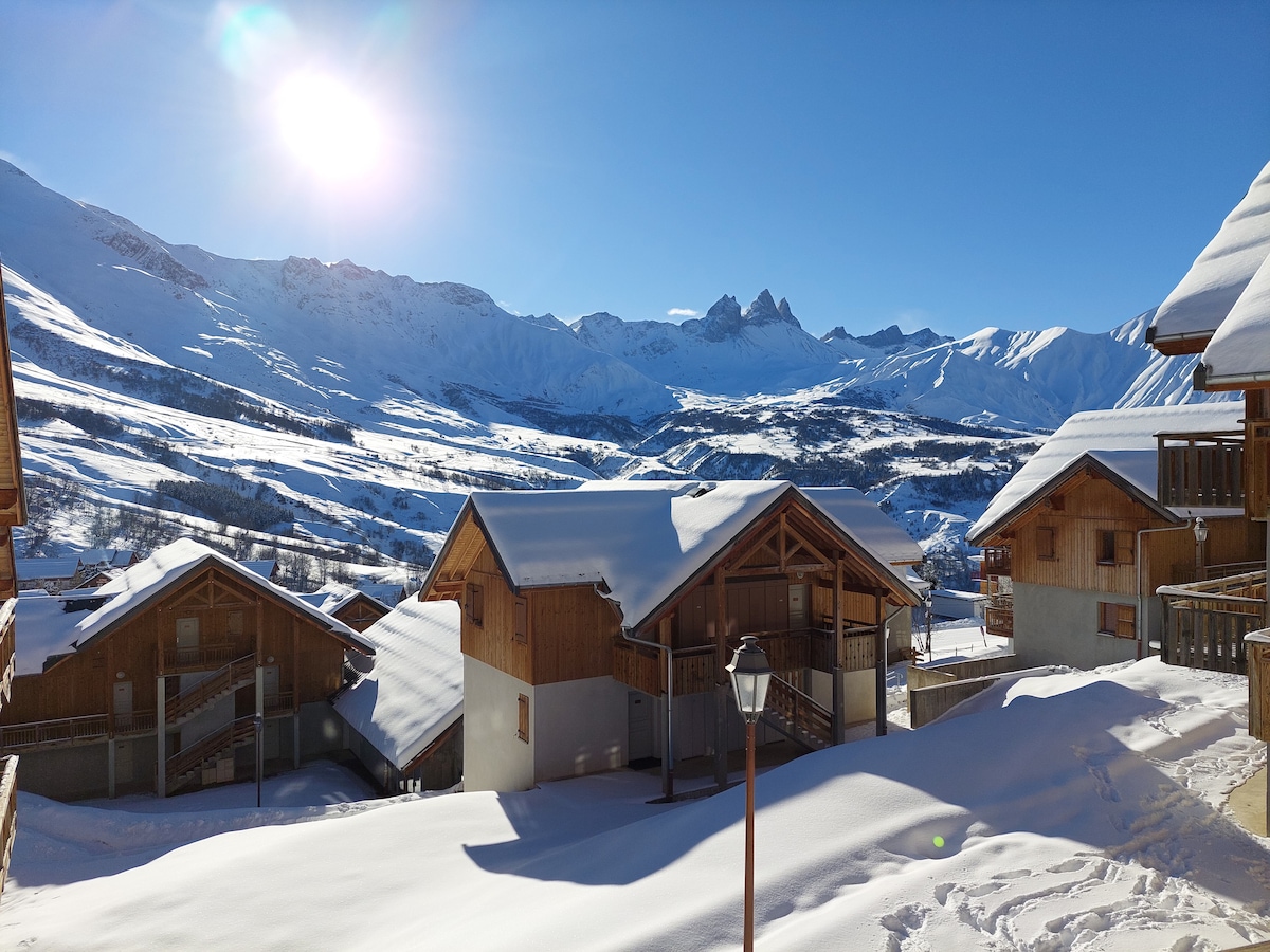 The image captures a bright winter landscape featuring wooden chalet-style buildings nestled amidst snow-covered slopes. Majestic mountains rise in the background under a clear blue sky, with the sun illuminating the scene, creating a serene atmosphere.