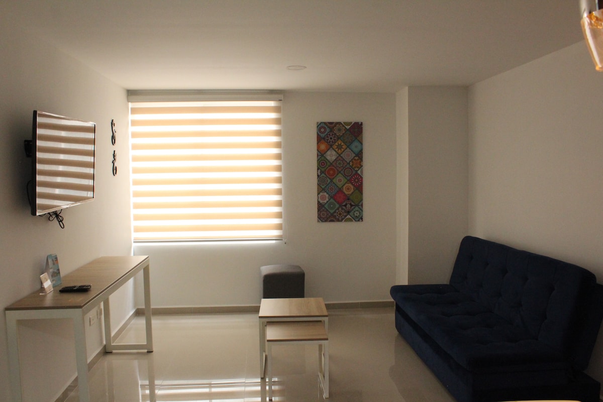 A well-lit living area is furnished with a dark blue sofa, a small coffee table, and a grey ottoman. A television is mounted on the wall opposite the seating. Natural light enters through the neatly arranged window with striped shades, highlighting the modern decor.