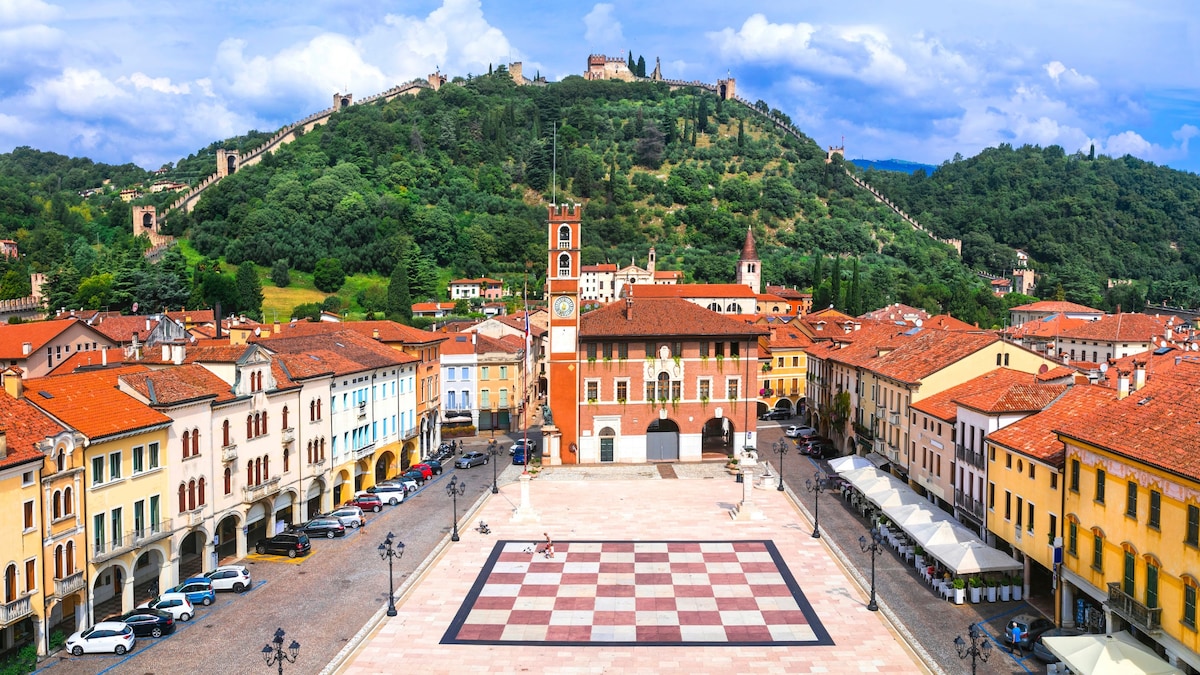 A panoramic view of a charming square is presented, featuring colorful buildings lining the edges. In the background, a hill with historic walls and greenery complements the scene, while the sky is dotted with clouds, creating a peaceful ambiance.
