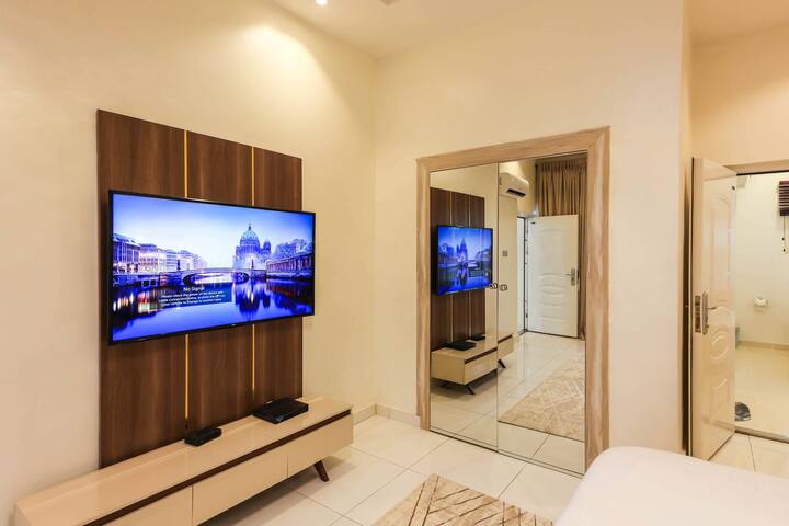 Sala de estar fresca y moderna con un televisor en la pared con un acento de madera, una bonita consola y una puerta espejada. Es un espacio luminoso y abierto con colores claros, pisos brillantes y cosas modernas, ideal para relajarse y tener amigos.