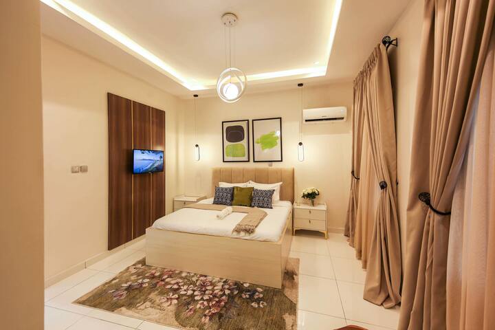 Dormitorio moderno: cama tamaño king, tonos neutros, detalles lujosos, asientos en las esquinas, iluminación ambiental, TV, cortinas exuberantes. Retiro sereno.