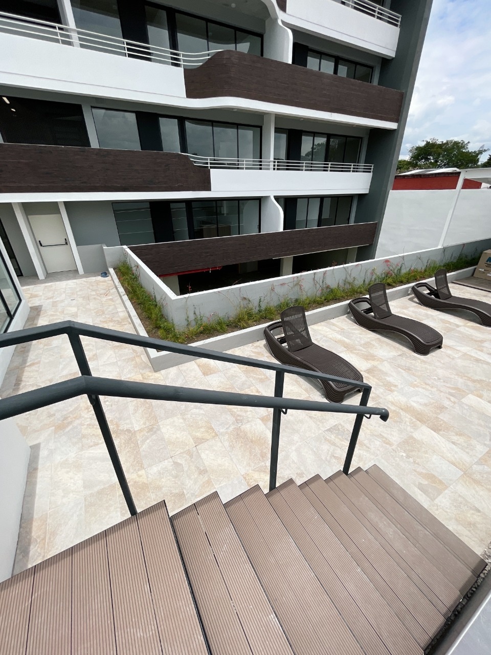 A set of stairs leads down to a spacious outdoor area featuring several lounge chairs arranged around a clean, tiled surface. The modern building's exterior looms above, showcasing a mix of dark and light materials with large windows and balconies.
