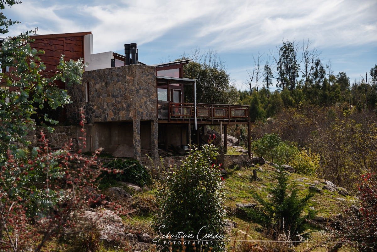 The image captures a spacious home set on a landscaped terrain, featuring a wooden deck that extends from the stone foundation. Surrounding greenery and natural rock formations contribute to the serene outdoor environment, while trees and shrubs frame the view.