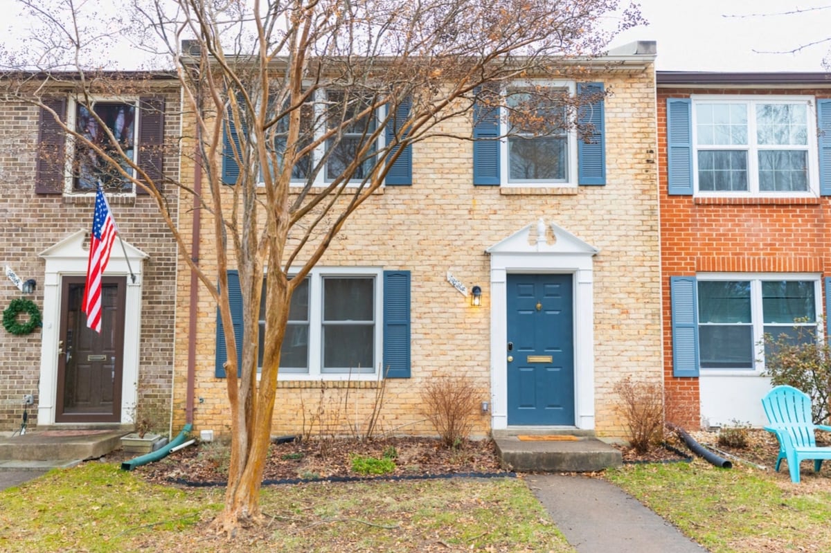 A townhouse with a brick and siding facade features a blue front door and white trim. The front yard is landscaped with small shrubs and a young tree. An Adirondack chair in light blue is positioned to the right, complementing the entryway.