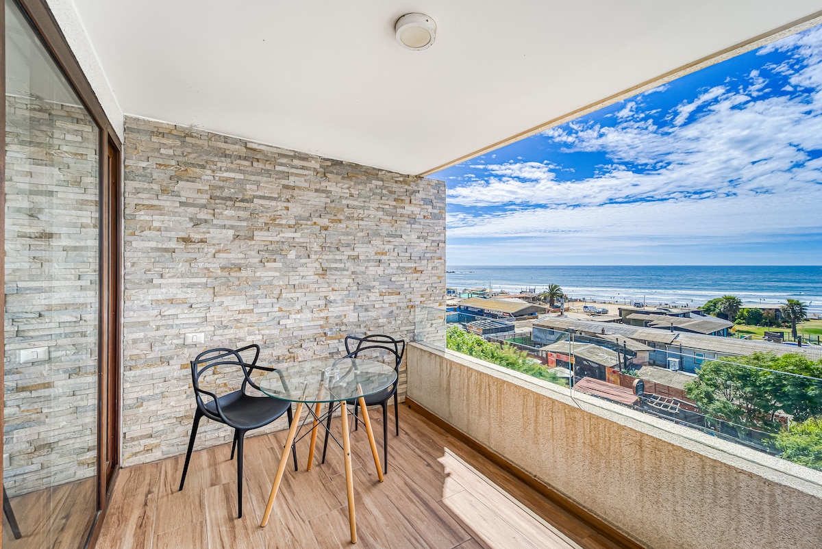 A terrace features a round glass table surrounded by two black chairs, offering a clear view of the coastline and ocean. The wall behind is styled with stone tiles, while large windows allow natural light to fill the space.