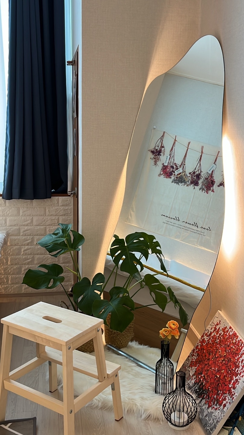A large, elegantly shaped mirror reflects light and creates an open ambiance. A wooden stool is positioned in front, with a backdrop of a potted plant for a touch of greenery. Decorative vases and wall art enhance the inviting atmosphere.