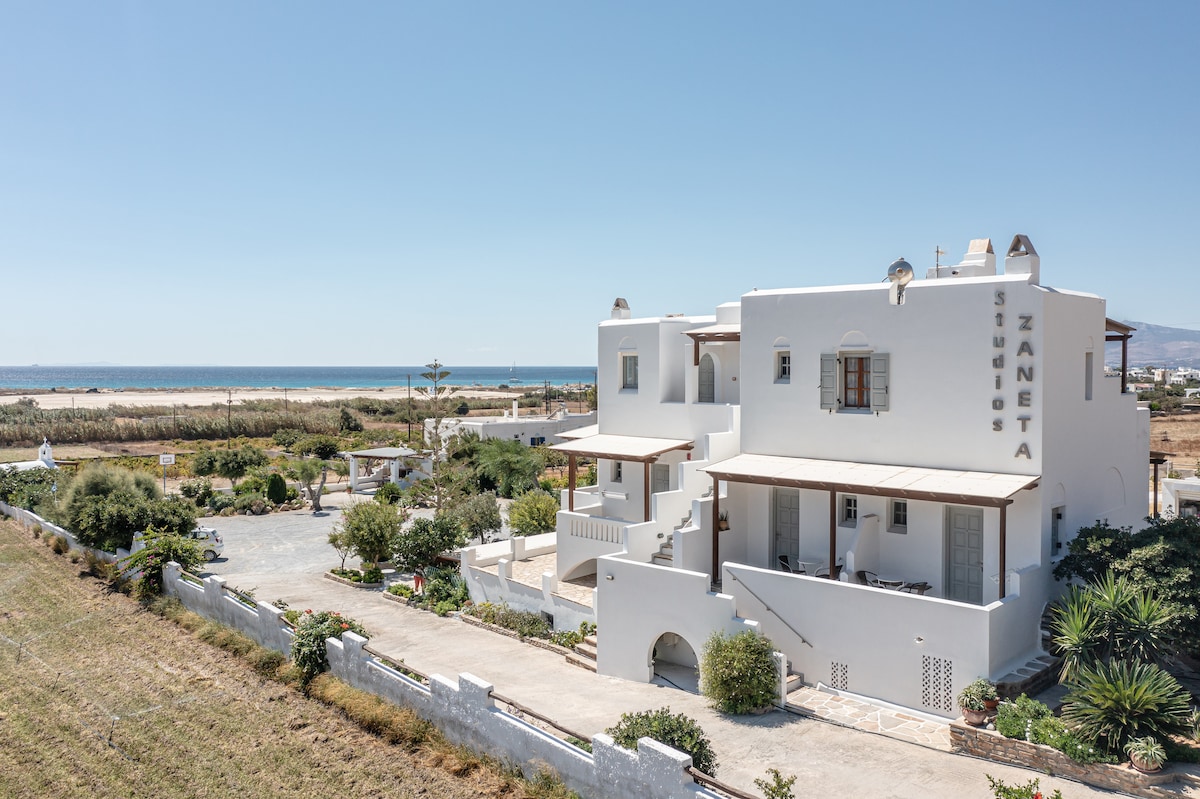 A white multi-level building with balconies overlooks lush greenery and a spacious parking area. A glimpse of the sea is visible in the background, enhancing the serene environment. The surrounding landscape features well-maintained gardens and open spaces.