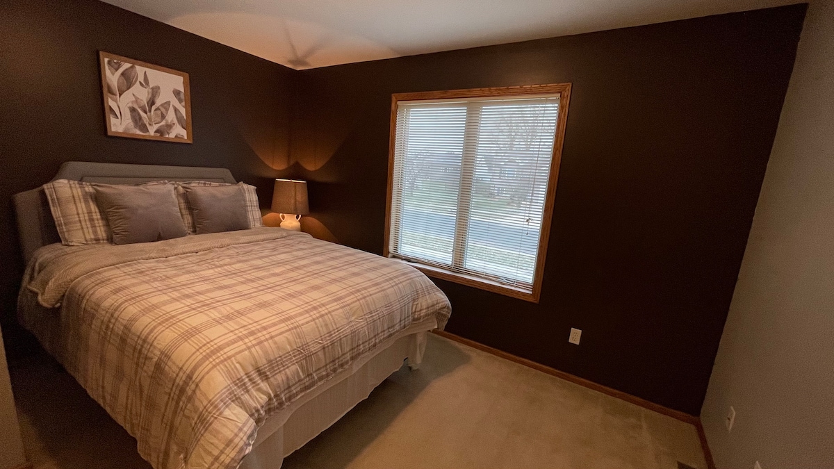 A well-appointed bedroom features a spacious bed with a soft plaid comforter and several pillows. Natural light enters through a large window, framed by blinds. A bedside lamp adds gentle illumination, and dark walls create a cozy ambiance.