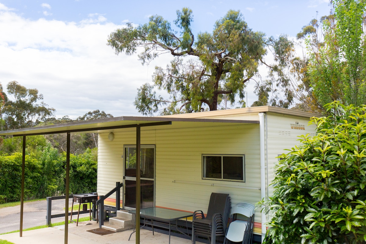 A cabin is situated within a lush green setting, featuring a shaded patio area. Outdoor seating is provided with several chairs and a small table. Surrounding trees contribute to the natural ambiance, complementing the simplicity of the structure.