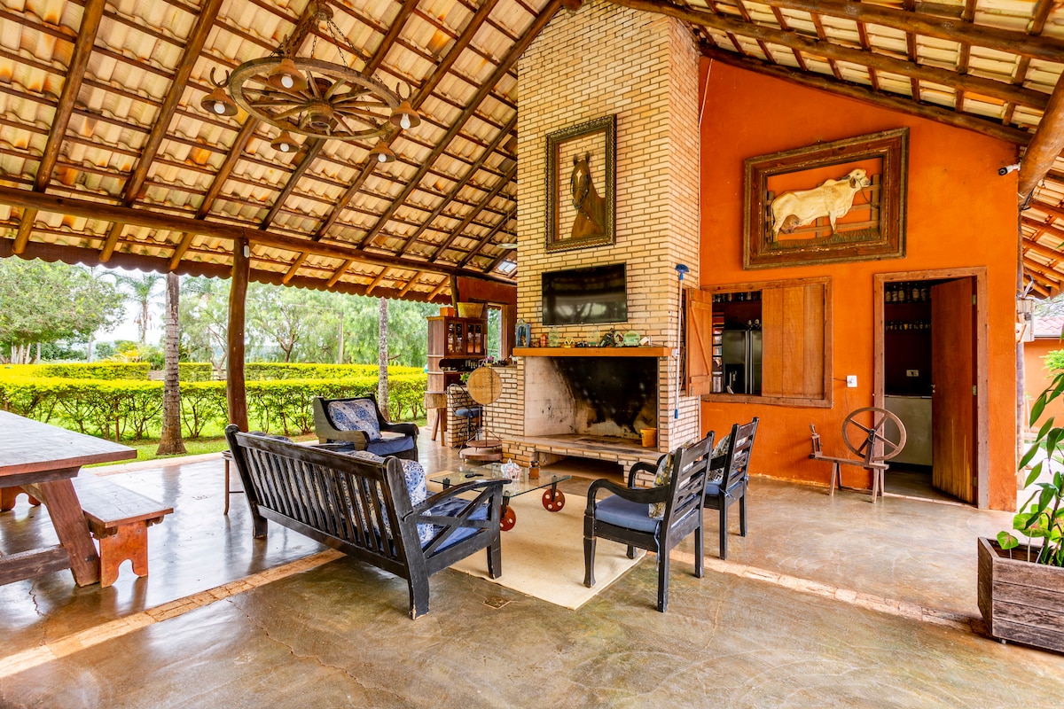 An open-air lounge area features wooden seating arrangements, with plush chairs surrounding a central fireplace. Decorative artworks adorn the rustic walls, while a thatched roof provides shade. Lush greenery is visible in the backdrop, enhancing the calm and inviting ambience.