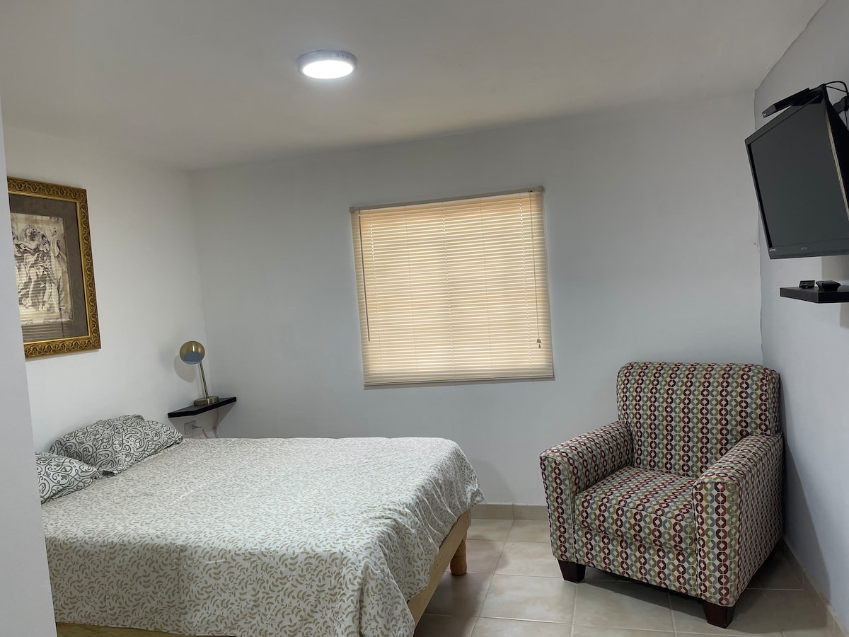 A spacious sleeping area features a bed with a patterned white and beige coverlet. A window with blinds allows soft light to enter. A patterned armchair is positioned nearby, with a lamp and a small shelf displaying a television mounted on the wall.