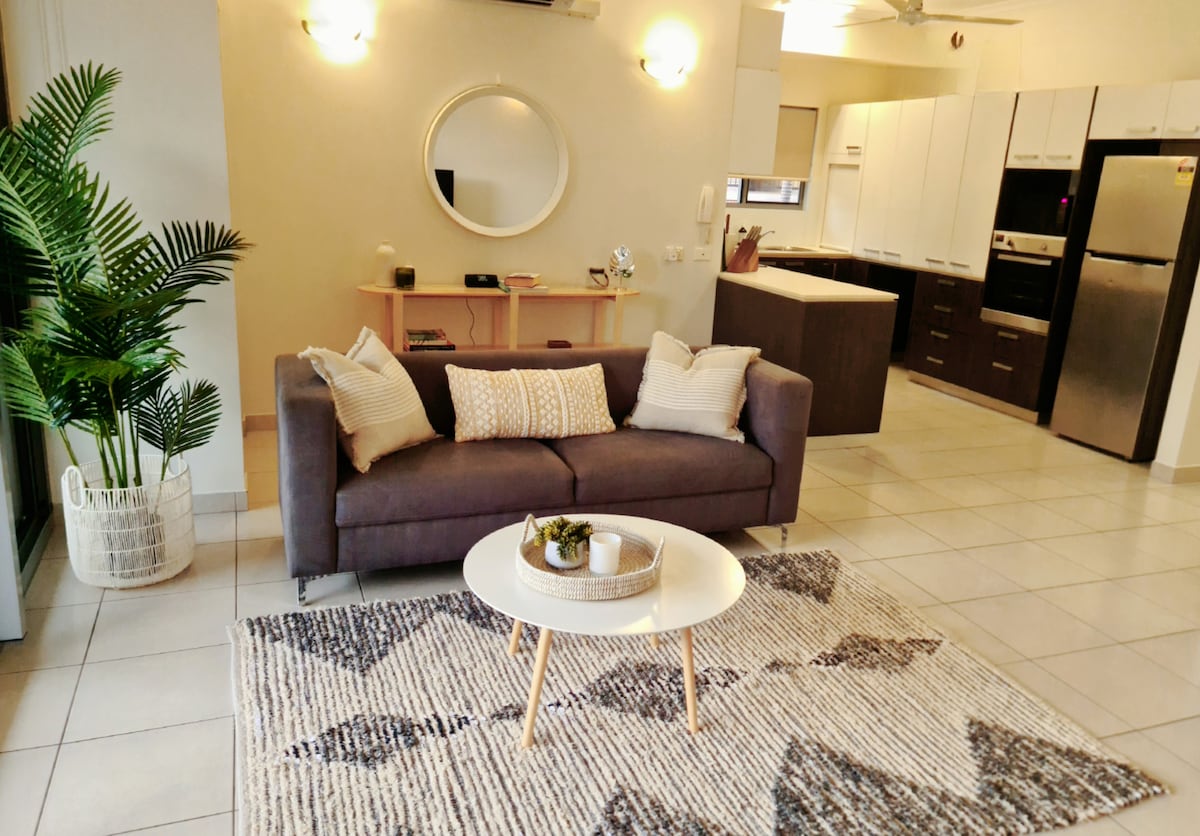 A modern living area features a comfortable dark sofa adorned with patterned cushions. A round coffee table sits on a textured rug, while a large indoor plant adds a touch of greenery. The open kitchen in the background is equipped with sleek cabinetry and appliances.