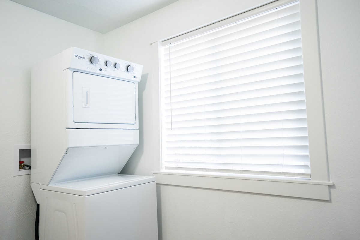 A laundry area features a stacked washer and dryer set against a bright white wall. Natural light filters through a window adorned with closed horizontal blinds, creating a well-lit and functional space.