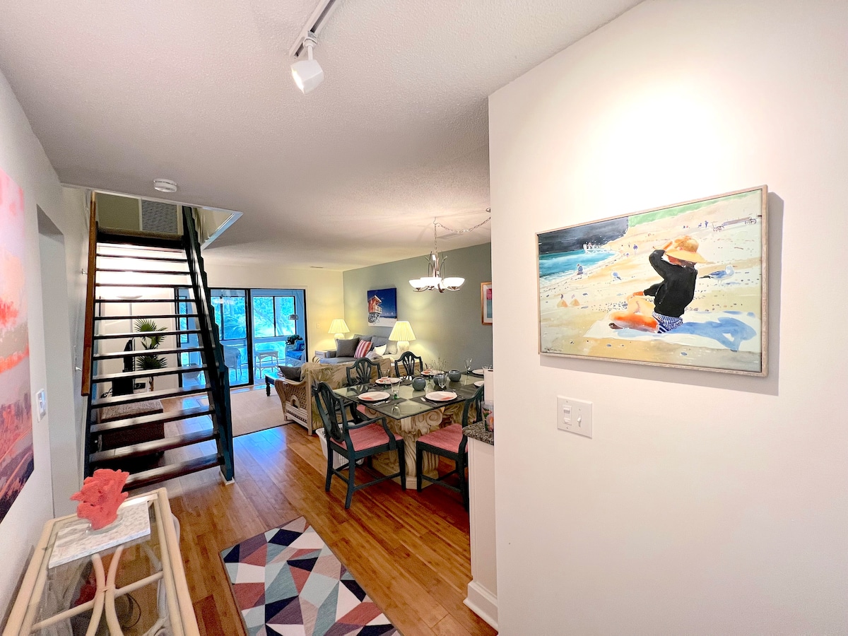 A view from the entrance showcases an open living area with seating arranged around a dining table. A staircase leads to the upper floor, while artwork hangs on the wall. Light colors create a bright atmosphere, complemented by natural light from a window.