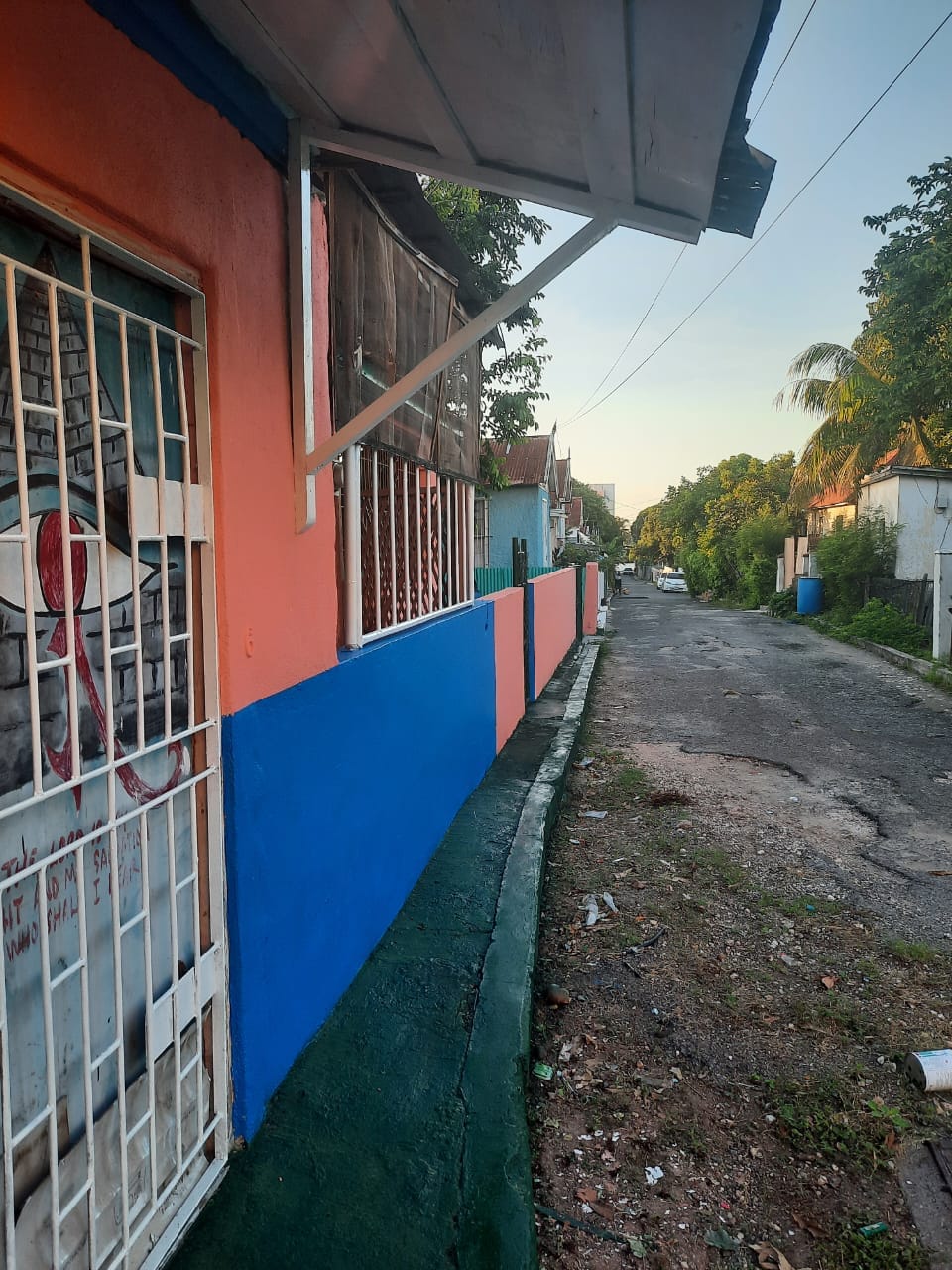 The image captures a colorful section of a neighborhood, featuring a building with vibrant orange and blue walls. A secured gate is visible in the foreground, while a quiet street extends into the background, lined with greenery and low buildings.