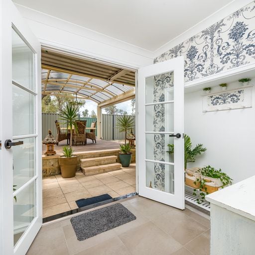A doorway leads to a spacious outdoor area featuring a covered patio. Potted plants and comfortable seating are visible, inviting relaxation. The interior is highlighted by decorative wallpaper and a soft grey mat at the entrance, creating a seamless transition from indoor to outdoor living.
