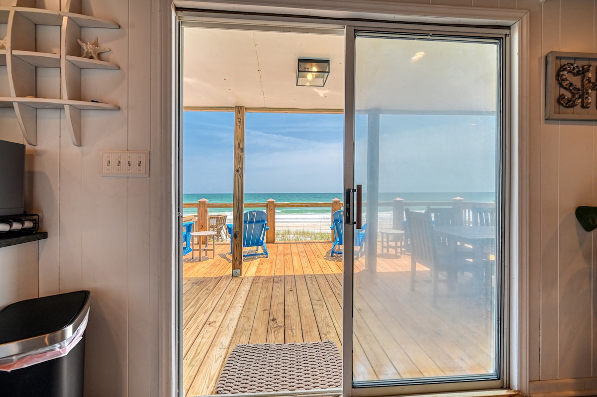 A sliding glass door provides a view of the spacious deck and ocean beyond. Light wood flooring on the deck complements outdoor furniture, including adirondack chairs and a dining table. The ocean waves can be seen gently rolling in the background, enhancing the serene atmosphere.