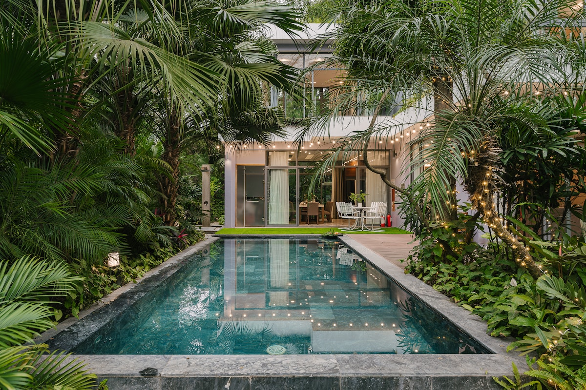 A serene private pool is surrounded by lush greenery and tropical plants, creating a tranquil outdoor space. Soft lighting is visible, enhancing the relaxing atmosphere. The villa is partially seen in the background, featuring large glass doors that connect it to the garden.