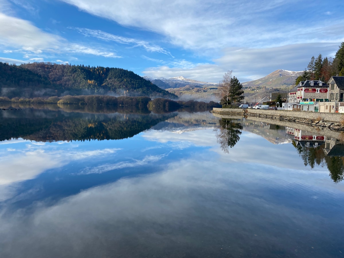 A serene lakeside view is presented, featuring calm waters that reflect the surrounding mountains and sky. The shoreline is lined with a mix of trees and buildings, providing a tranquil setting for leisurely walks along the water's edge.