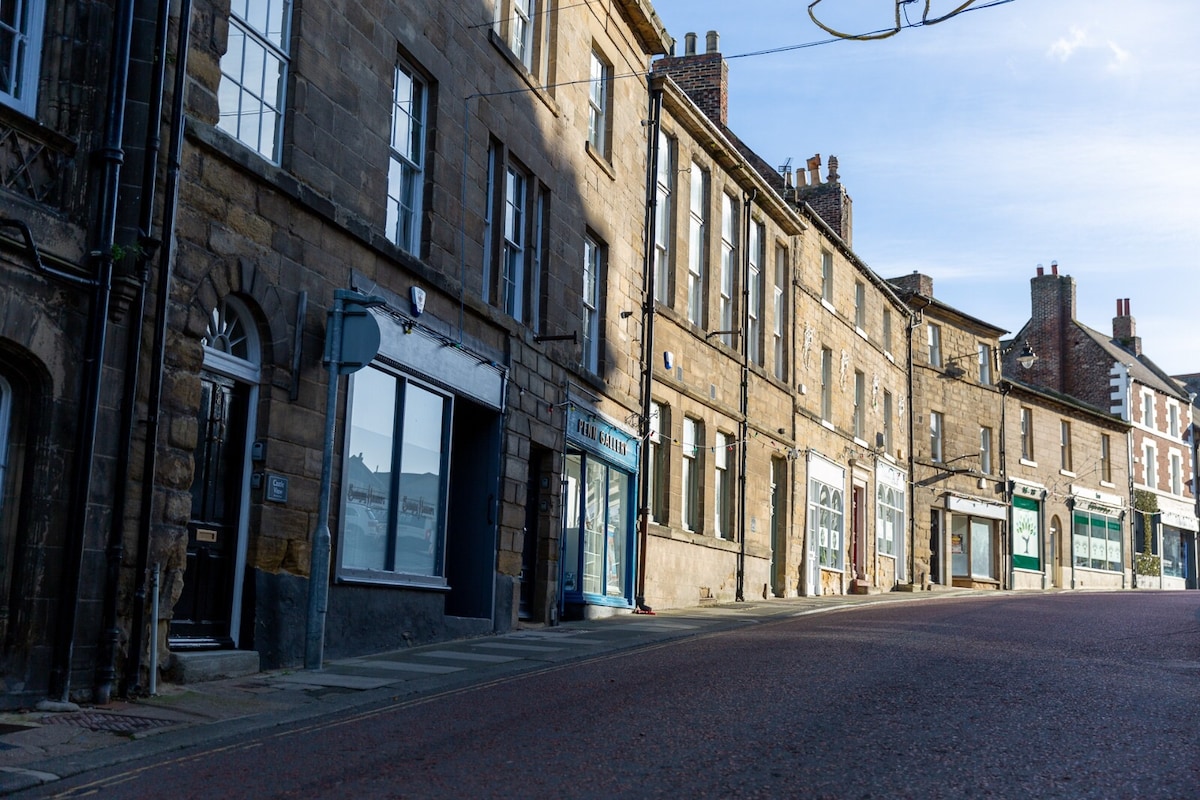 A quiet street in a historic market town is lined with stone buildings featuring large windows. The buildings showcase various facades and styles, with some displaying signs. A gently sloping road leads the eye toward the horizon under a clear blue sky.