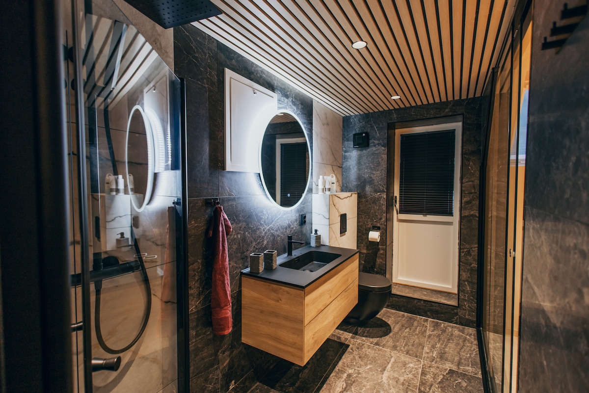 A modern bathroom is displayed, featuring a large, illuminated round mirror above a wooden sink cabinet. A walk-in shower is visible on one side, with sleek tiles covering the walls and floor. A large window offers natural light, while a frosted glass panel can be seen.