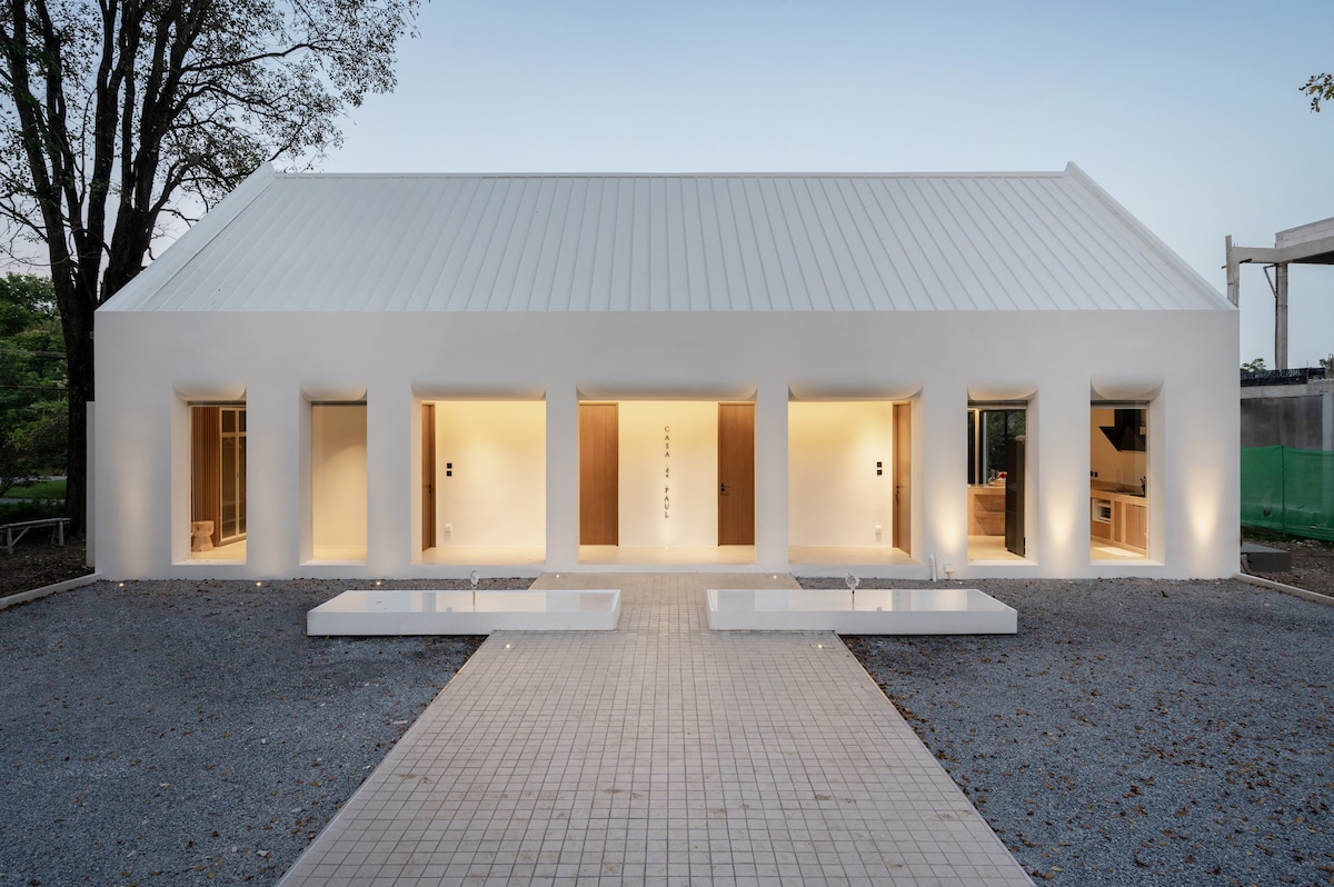 The exterior of the house features a modern, minimalist design with a clean white facade. Large, rectangular openings provide indirect lighting, enhancing the architectural lines. A pathway lined with stone tiles leads to the entrance, and surrounding greenery offers a natural contrast.