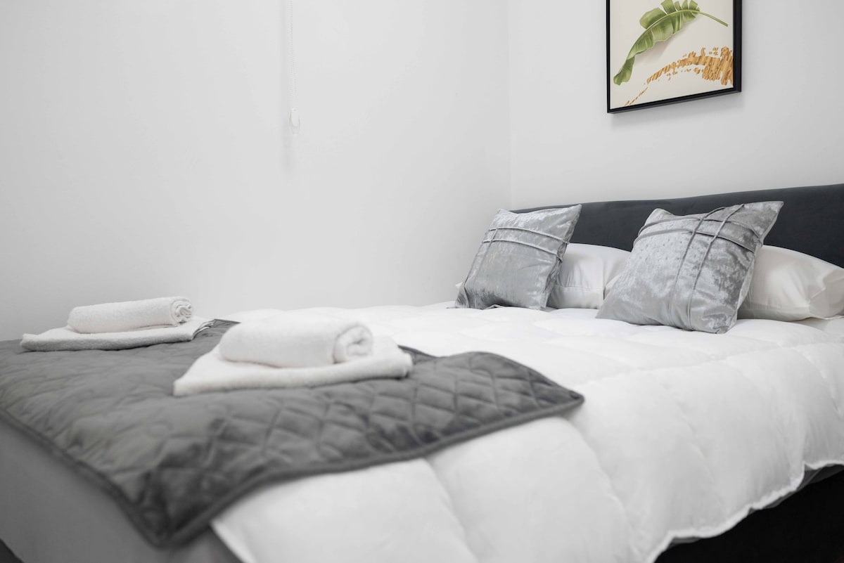 A neatly made bed showcases plush pillows and a decorative throw. Soft white linens are complemented by a silver quilt. A framed botanical print hangs on the wall above, adding a touch of elegance to the serene space.