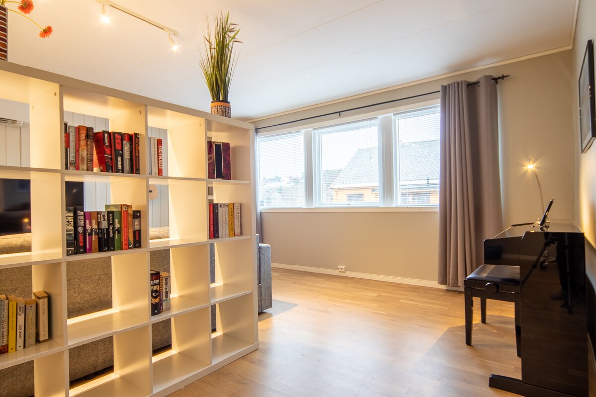 A bright living area is defined by a modern bookshelf filled with various books, separating it from a corner with a piano. Large windows allow natural light to fill the space, accented by neutral wall tones and light hardwood floors.