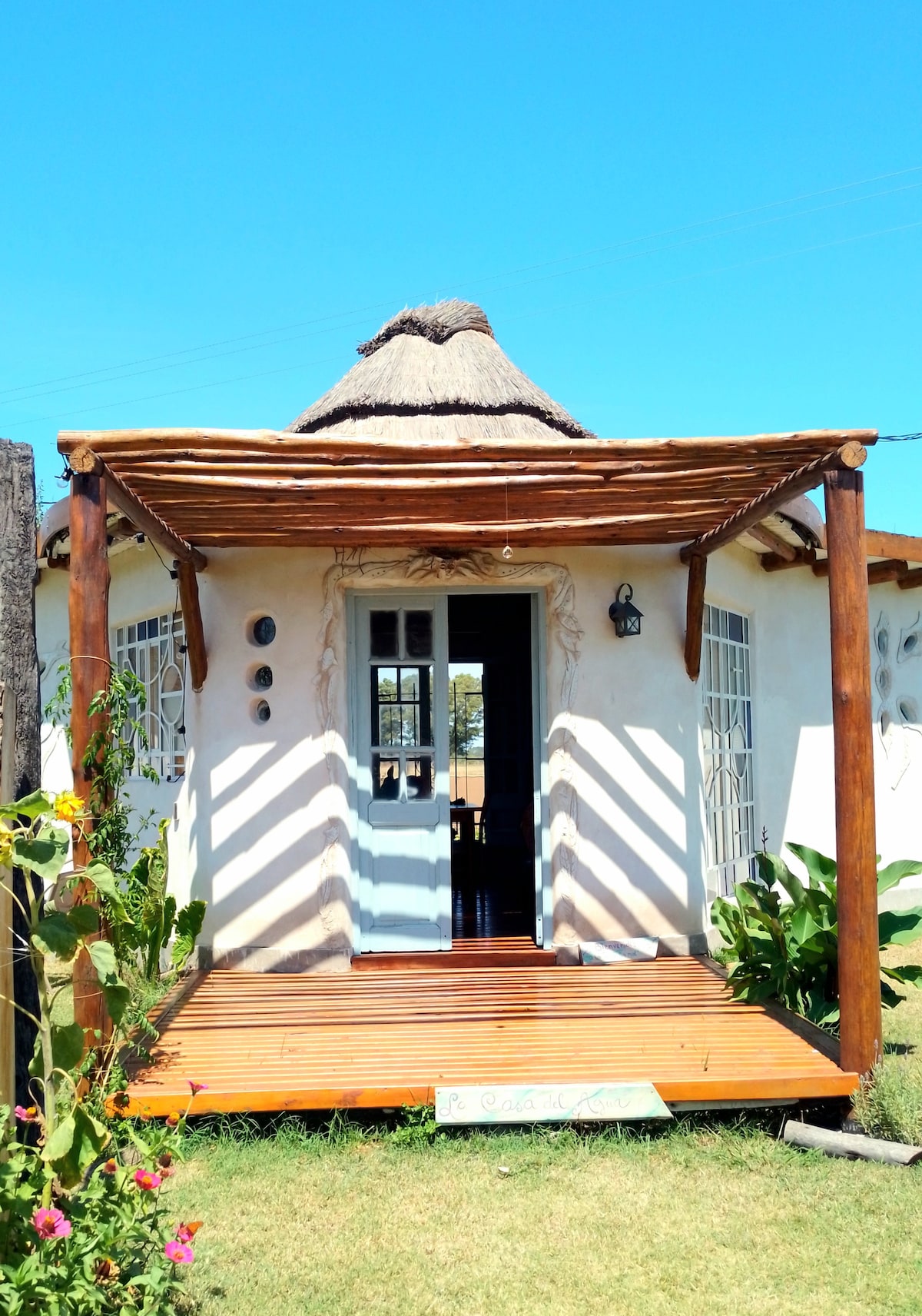 The entrance features a wooden deck with a thatched roof overhead, providing shade. Light-colored walls are complemented by white-framed windows, and decorative elements add character. Lush greenery surrounds the entrance, enhancing the welcoming atmosphere.