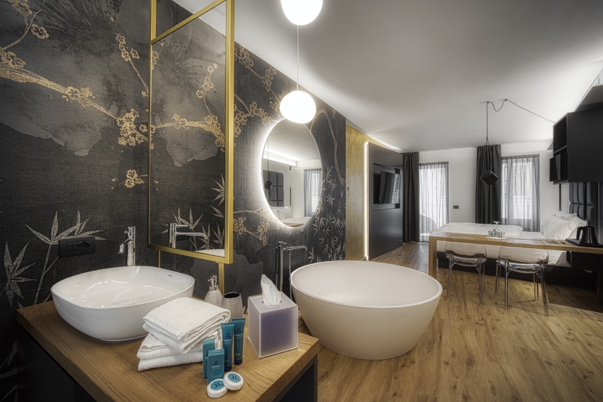 An open living space is highlighted by a modern round bathtub and a stylish sink with a large mirror above. Light-colored wooden flooring complements the artistic wall design. A spacious bed and a TV are positioned in the background, with natural light filtering through the windows.
