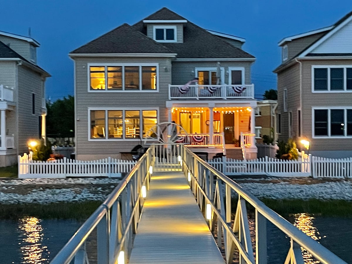 The large three-story waterfront home is illuminated at dusk, showcasing bright windows and a welcoming front porch adorned with festive decorations. A well-lit walkway extends over the water to the house, bordered by a white fence, providing easy access for guests.