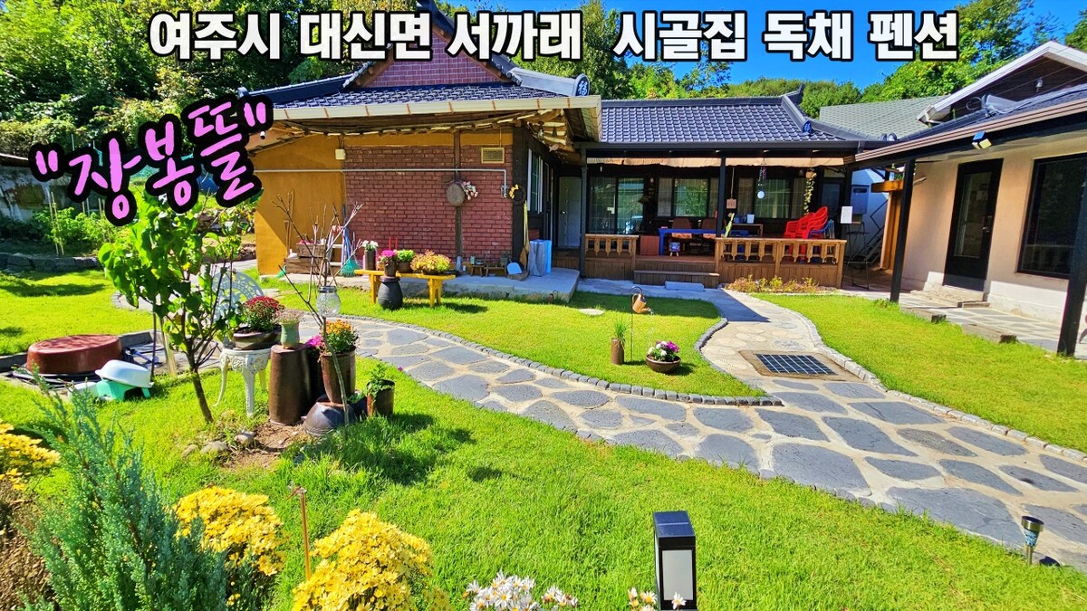 A serene garden surrounds a traditional style house, featuring a stone pathway that leads to a cozy outdoor seating area. Lush green grass, flowering plants, and decorative lighting create a tranquil setting, ideal for relaxation away from urban life.